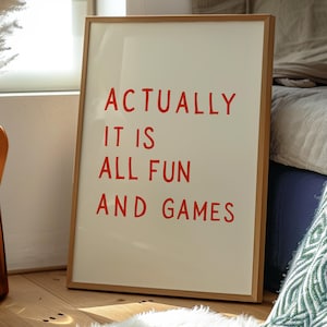 May include: A framed art print with the words "ACTUALLY IT IS ALL FUN AND GAMES" in red letters. The artwork is in a light wood frame and leans against a wall. The background includes a fluffy white rug and a bed.