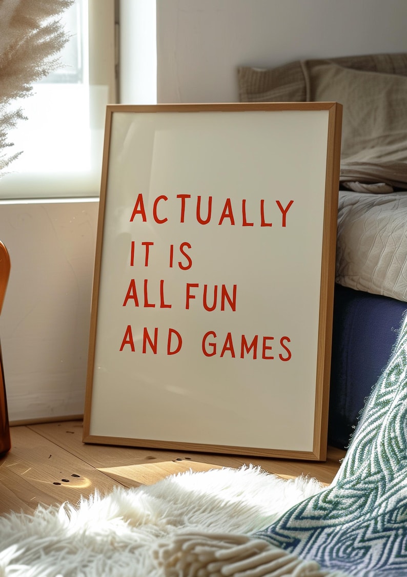 May include: A framed art print with the words "ACTUALLY IT IS ALL FUN AND GAMES" in red letters. The artwork is in a light wood frame and leans against a wall. The background includes a fluffy white rug and a bed.