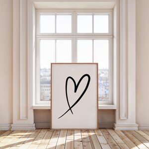 May include: A framed art print featuring a black heart drawn with a single brushstroke on a white background. The artwork is displayed in a room with a window and wooden flooring. The frame is a light brown color.