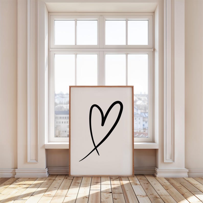 May include: A framed art print featuring a black heart drawn with a single brushstroke on a white background. The artwork is displayed in a room with a window and wooden flooring. The frame is a light brown color.