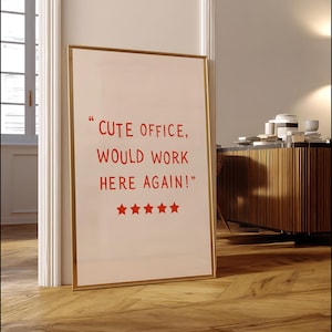 May include: A framed print with the text "CUTE OFFICE, WOULD WORK HERE AGAIN!" in red, along with five red stars. The print has a gold frame and is leaning against a white door frame on a wooden floor.