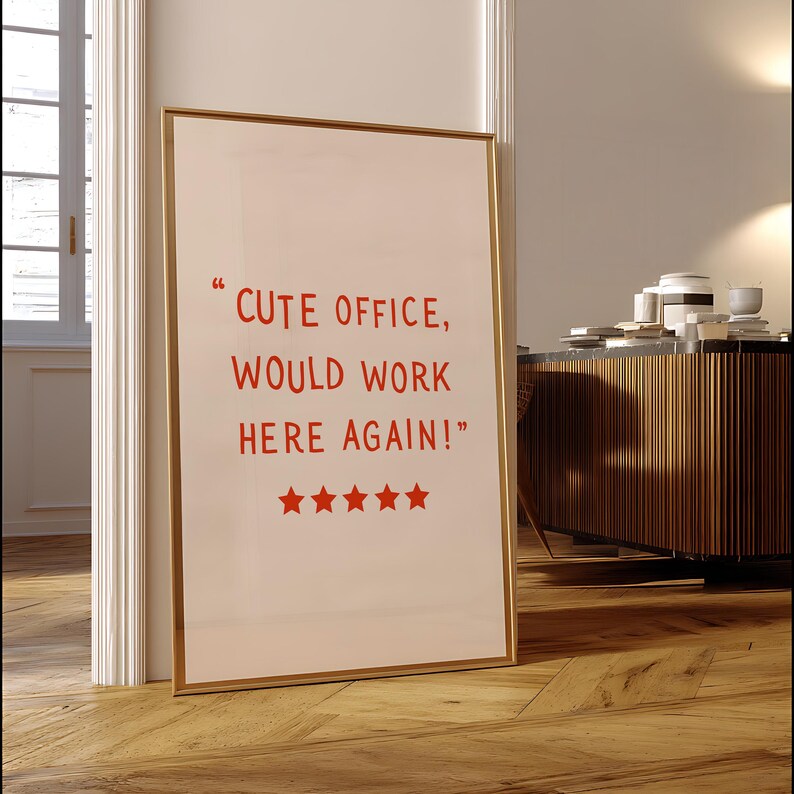 May include: A framed print with the text "CUTE OFFICE, WOULD WORK HERE AGAIN!" in red, along with five red stars. The print has a gold frame and is leaning against a white door frame on a wooden floor.