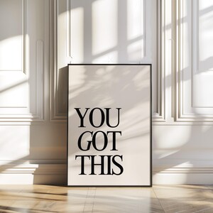 K&ouml;nnte beinhalten: Gerahmter Druck mit den Worten "YOU GOT THIS" in fetten schwarzen Buchstaben auf cremefarbenem Hintergrund. Der Druck befindet sich in einem schwarzen Rahmen und lehnt an einer wei&szlig;en Wand mit Leisten auf einem Holzboden.
