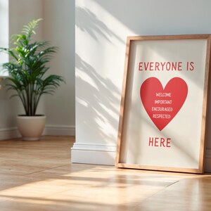 May include: A framed print with the words "EVERYONE IS HERE" in red, with a red heart graphic. The heart contains the words "WELCOME, IMPORTANT, ENCOURAGED, RESPECTED". The frame is wood-toned, and the print leans against a white wall.