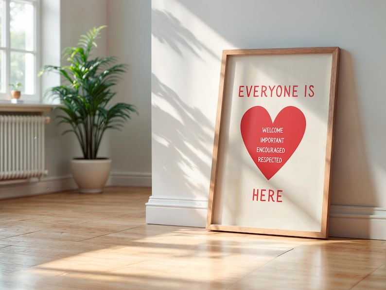 May include: A framed print with the words "EVERYONE IS HERE" in red, with a red heart graphic. The heart contains the words "WELCOME, IMPORTANT, ENCOURAGED, RESPECTED". The frame is wood-toned, and the print leans against a white wall.