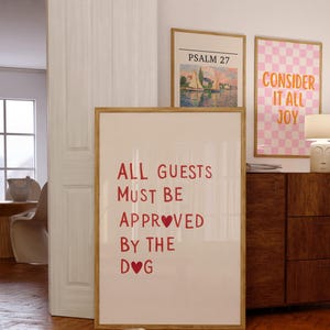 Könnte beinhalten: Ein gerahmter Druck mit dem Text "ALL GUESTS MUST BE APPROVED BY THE DOG" in roten Buchstaben. Zwei weitere gerahmte Drucke sind sichtbar, einer mit einem Landschaftsgemälde und der andere mit einem rosa-weiß karierten Muster und den Worten "CONSIDER IT ALL JOY".