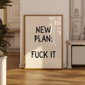 以下が含まれることがあります： 「NEW PLAN: FUCK IT」のテキストが黒で、クリーム色の背景に中央に配置されたフレーム付きプリント。フレームは細いゴールド色です。プリントは木製の床に表示されています。