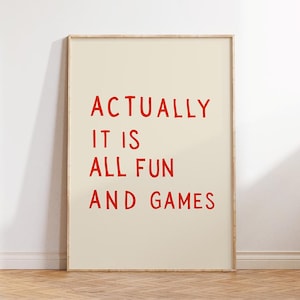 May include: A framed art print with the words "ACTUALLY IT IS ALL FUN AND GAMES" in red, bold letters on a cream-colored background. The artwork is displayed in a light-colored wooden frame.