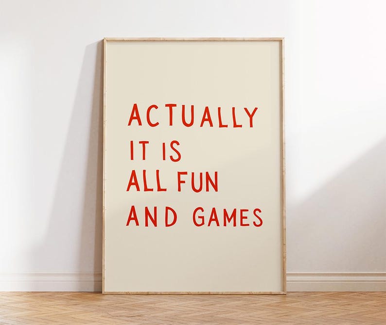May include: A framed art print with the words "ACTUALLY IT IS ALL FUN AND GAMES" in red, bold letters on a cream-colored background. The artwork is displayed in a light-colored wooden frame.