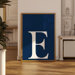 May include: A framed art print featuring a large, white letter "E" against a deep navy blue background. The artwork is encased in a gold-colored frame, creating a striking contrast. The print is displayed in a well-lit room with hardwood flooring.