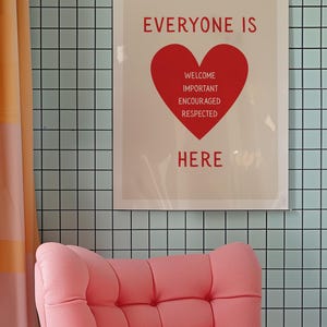 May include: A framed poster with the words "EVERYONE IS HERE" in red, with a red heart graphic. Below the heart are the words "WELCOME, IMPORTANT, ENCOURAGED, RESPECTED." A pink armchair is in the foreground, and the background is a light blue tiled wall.