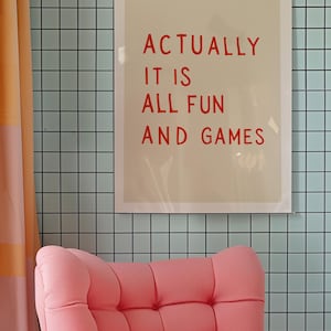 May include: A framed print with the text "ACTUALLY IT IS ALL FUN AND GAMES" in red letters hangs on a light blue tiled wall. A pink armchair sits below the print, and a peach-colored curtain is on the left.
