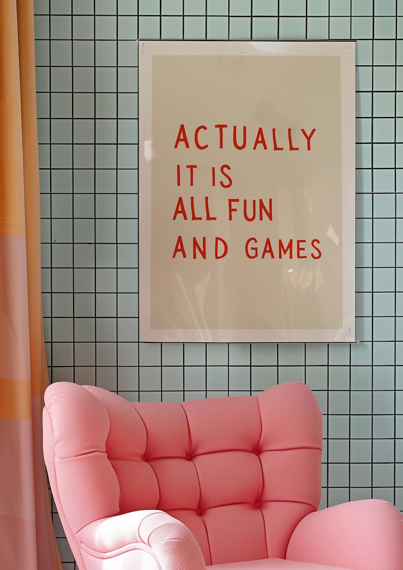 May include: A framed print with the text "ACTUALLY IT IS ALL FUN AND GAMES" in red letters hangs on a light blue tiled wall. A pink armchair sits below the print, and a peach-colored curtain is on the left.