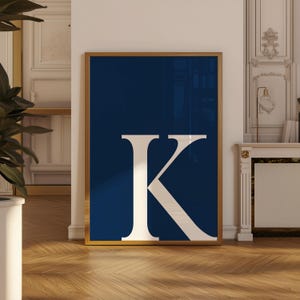 May include: A framed art print featuring a large white letter "K" against a deep navy blue background. The artwork is encased in a gold-colored frame and leans against a white wall. The floor is made of wood.