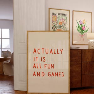 May include: A framed poster with the text "ACTUALLY IT IS ALL FUN AND GAMES" in red. Two other framed art pieces are visible, one with floral art and the other with a bouquet of tulips. The frames are a light brown color.