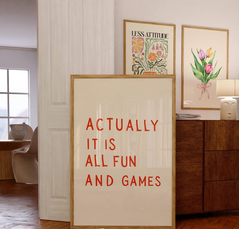 May include: A framed poster with the text "ACTUALLY IT IS ALL FUN AND GAMES" in red. Two other framed art pieces are visible, one with floral art and the other with a bouquet of tulips. The frames are a light brown color.