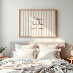 May include: A bedroom scene featuring a bed with cream-colored bedding and pillows. Above the bed is a framed artwork with vertical stripes and the text "how lucky are we". A small side table holds a plant.