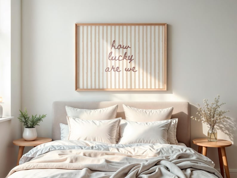 May include: A bedroom scene featuring a bed with cream-colored bedding and pillows. Above the bed is a framed artwork with vertical stripes and the text "how lucky are we". A small side table holds a plant.