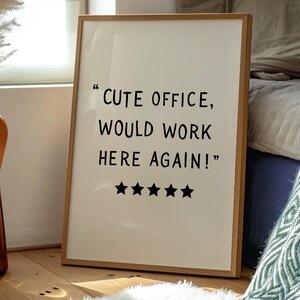 May include: A framed print with the text "CUTE OFFICE, WOULD WORK HERE AGAIN!" and five black stars. The frame is light brown, and the background is off-white. The print is leaning against a wall.
