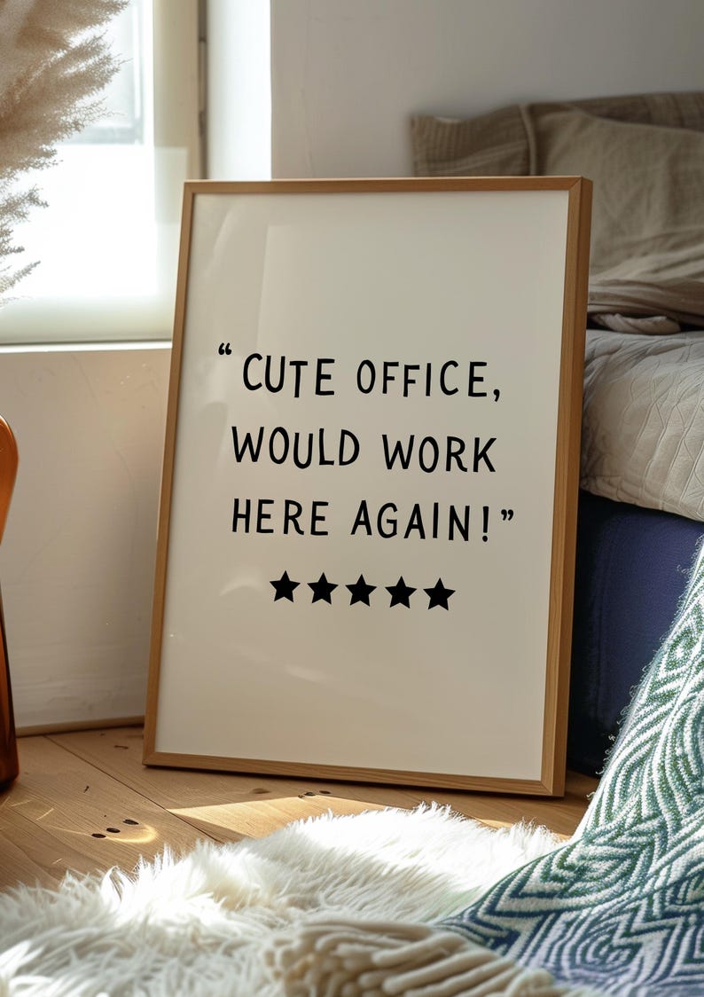 May include: A framed print with the text "CUTE OFFICE, WOULD WORK HERE AGAIN!" and five black stars. The frame is light brown, and the background is off-white. The print is leaning against a wall.