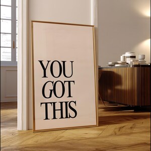 K&ouml;nnte beinhalten: Ein gerahmter Druck mit dem Text "YOU GOT THIS" in fetten schwarzen Buchstaben. Der Druck hat einen goldenen Rahmen und lehnt an einem wei&szlig;en T&uuml;rrahmen auf einem Holzboden. Im Hintergrund befindet sich ein Holzschrank.