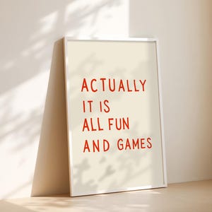 May include: A framed print with the text "ACTUALLY IT IS ALL FUN AND GAMES" in red letters on an off-white background. The white frame leans against a wall, casting a shadow.