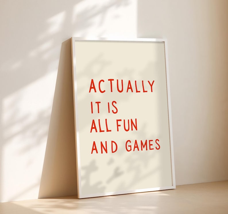 May include: A framed print with the text "ACTUALLY IT IS ALL FUN AND GAMES" in red letters on an off-white background. The white frame leans against a wall, casting a shadow.