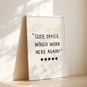 May include: A framed print with the text "CUTE OFFICE, WOULD WORK HERE AGAIN!" in black font. Below the text are four black stars. The frame is white, and the background is a light beige color.