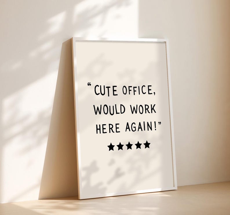 May include: A framed print with the text "CUTE OFFICE, WOULD WORK HERE AGAIN!" in black font. Below the text are four black stars. The frame is white, and the background is a light beige color.