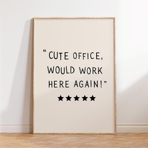 May include: A framed print with the text "CUTE OFFICE, WOULD WORK HERE AGAIN!" in black font. Below the text are five black star icons. The background is a light beige color, and the frame is a light wood color.