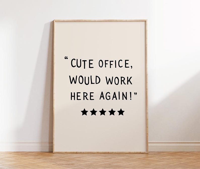 May include: A framed print with the text "CUTE OFFICE, WOULD WORK HERE AGAIN!" in black font. Below the text are five black star icons. The background is a light beige color, and the frame is a light wood color.