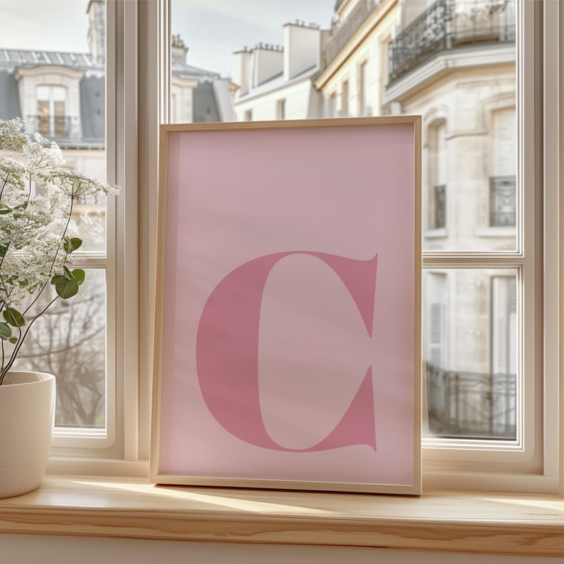 Puede incluir: Un p&oacute;ster rosa enmarcado con una gran letra rosa "C" en el centro. El p&oacute;ster est&aacute; colgado en una pared junto a una ventana con vista a una ciudad.