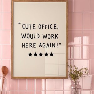 May include: A framed print with the text "CUTE OFFICE, WOULD WORK HERE AGAIN!" in black font. Below the text are five black stars. The frame is wood-toned, set against a pink tiled wall.