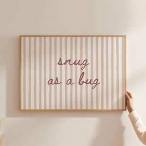 以下が含まれることがあります： ベージュと白のストライプの背景を持つ額入りの壁の芸術作品。 「snug as a bug」というテキストが筆記体でアートワークの中央に書かれています。 フレームは明るい茶色の木材で作られています。 手がアートワークを持っています。