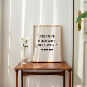 May include: A framed print with the text "Cute office, would work here again!" and five stars. The print is on a wooden table with a vase of white flowers. The room has a neutral color palette.