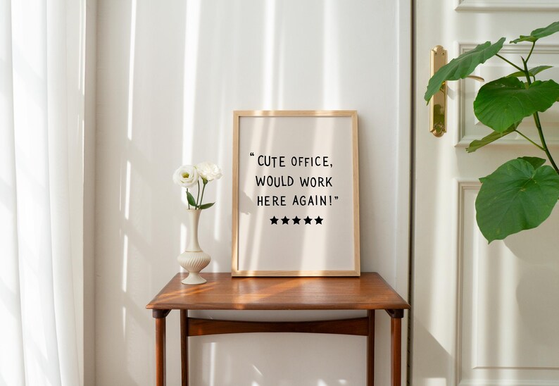 May include: A framed print with the text "Cute office, would work here again!" and five stars. The print is on a wooden table with a vase of white flowers. The room has a neutral color palette.