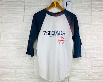 May include: A white and blue baseball t-shirt with a faded tie-dye effect on the blue sleeve. The shirt has a graphic print of "7 Seconds Japan Tour 2001" with a red circle and a cross inside.