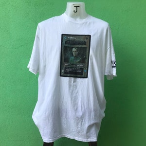May include: White t-shirt with a graphic of a card from the game "Dead Man's Hand". The card features a man in a suit with the text "Dead Man's Hand" above him.