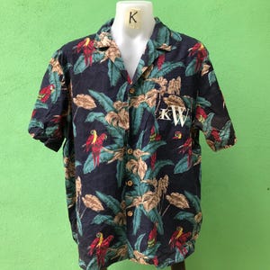 May include: Navy blue short-sleeved button-down shirt with a tropical parrot and floral print. The shirt features a classic collar, a chest pocket with the initials "KW", and wooden buttons. The shirt is displayed on a mannequin.