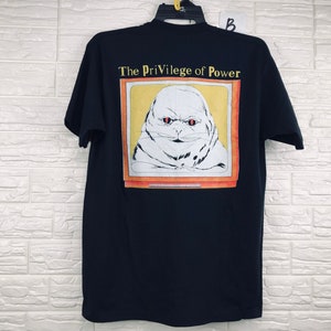 Könnte beinhalten: Schwarzes T-Shirt mit einem Grafikdruck eines Comic-Charakters mit roten Augen und gelbem Hintergrund. Der Text "The Privilege of Power" ist über dem Grafikdruck gedruckt.