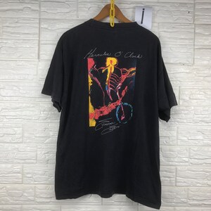 Puede incluir: Camiseta negra con un gráfico colorido de una persona montando en bicicleta. El gráfico está rodeado por un borde amarillo y el texto "Hercules O'Clock" está escrito encima. El texto "Guitartown" está escrito debajo del gráfico.