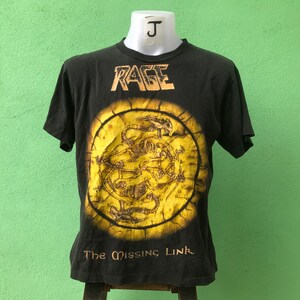 Puede incluir: Camiseta negra con la palabra "RAGE" en dorado sobre un gráfico circular. El gráfico tiene un fondo amarillo con un diseño detallado y el texto "The Missing Link" debajo.