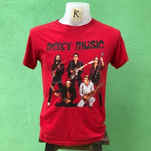 Könnte beinhalten: Rotes T-Shirt mit einem Band-Motiv von Roxy Music. Das Motiv zeigt fünf Bandmitglieder, die Instrumente spielen. Der Name der Band ist in schwarzen Buchstaben über dem Motiv gedruckt.