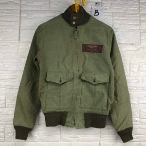 May include: A green bomber jacket with a brown patch that says "Eagle's Wings" on the left side of the chest. The jacket has two pockets on the front and a zipper closure.