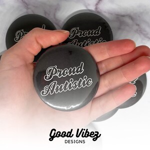 May include: Several black, circular badges with the white text "Proud Autistic" in a cursive font. The badges have a glossy finish. The words "Good Vibez Designs" are printed in a script font at the bottom of the image.