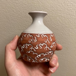 May include: A small, handcrafted ceramic vase with a rounded body and a narrow neck. The vase features a reddish-brown base adorned with white, swirling patterns. The neck is a contrasting off-white color, creating a two-tone effect.