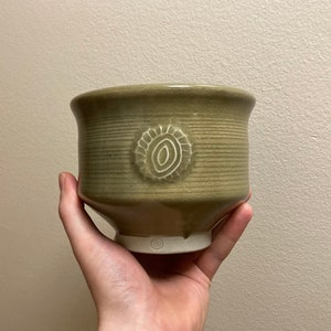 Könnte beinhalten: Eine grüne Keramikschüssel mit einem weißen Design auf der Vorderseite. Die Schüssel hat einen leicht ausgestellten Rand und einen abgerundeten Boden.