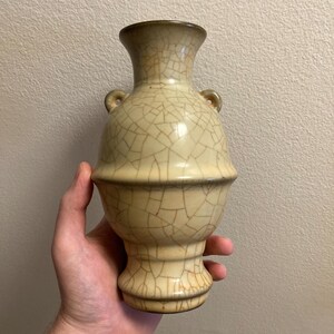 Könnte beinhalten: Eine beige Keramikvase mit einem Craquelé-Glasurmuster. Die Vase hat einen bauchigen Körper, einen ausgestellten Fuß und einen schmalen Hals mit zwei kleinen Schlaufengriffen. Die Vase wird in einer Hand gehalten.
