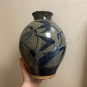 May include: A ceramic vase with a rounded body and a narrow neck. The vase is grey-green with dark blue leaf patterns. The base is a darker blue, and the bottom has a cork base.