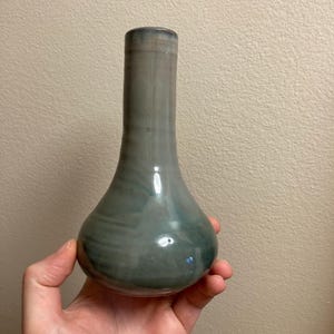 May include: A handcrafted, glazed ceramic vase with a bulbous base and a slender neck. The vase features a blend of gray and teal hues, with subtle marbling effects. The smooth, glossy finish reflects light, highlighting its elegant form.